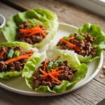 Freshly prepared Ground Beef Lettuce Wraps Recipe on a white ceramic plate, featuring crisp lettuce, sautéed ground beef, shredded carrots, and green onions, garnished with sriracha sauce and chopped peanuts. A healthy and low-carb meal option.